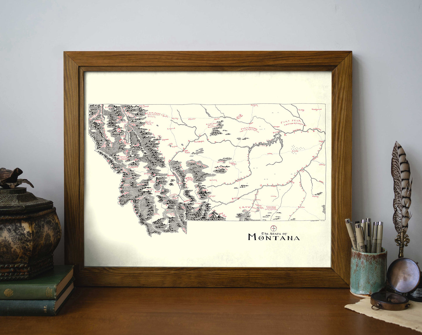 The Montana Map by Lord of Maps is a framed, vintage-style print featuring hand-drawn mountains, rivers, and towns. Displayed on a wooden desk with books, a decorative container, feather quill, and cup of pens and brushes nearby.