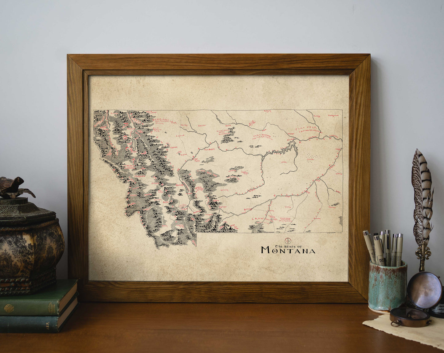 The Montana Map by Lord of Maps, a framed vintage-style print, is displayed on a wooden desk with books, a decorative box, a feather quill, and a container for pens and pencils.