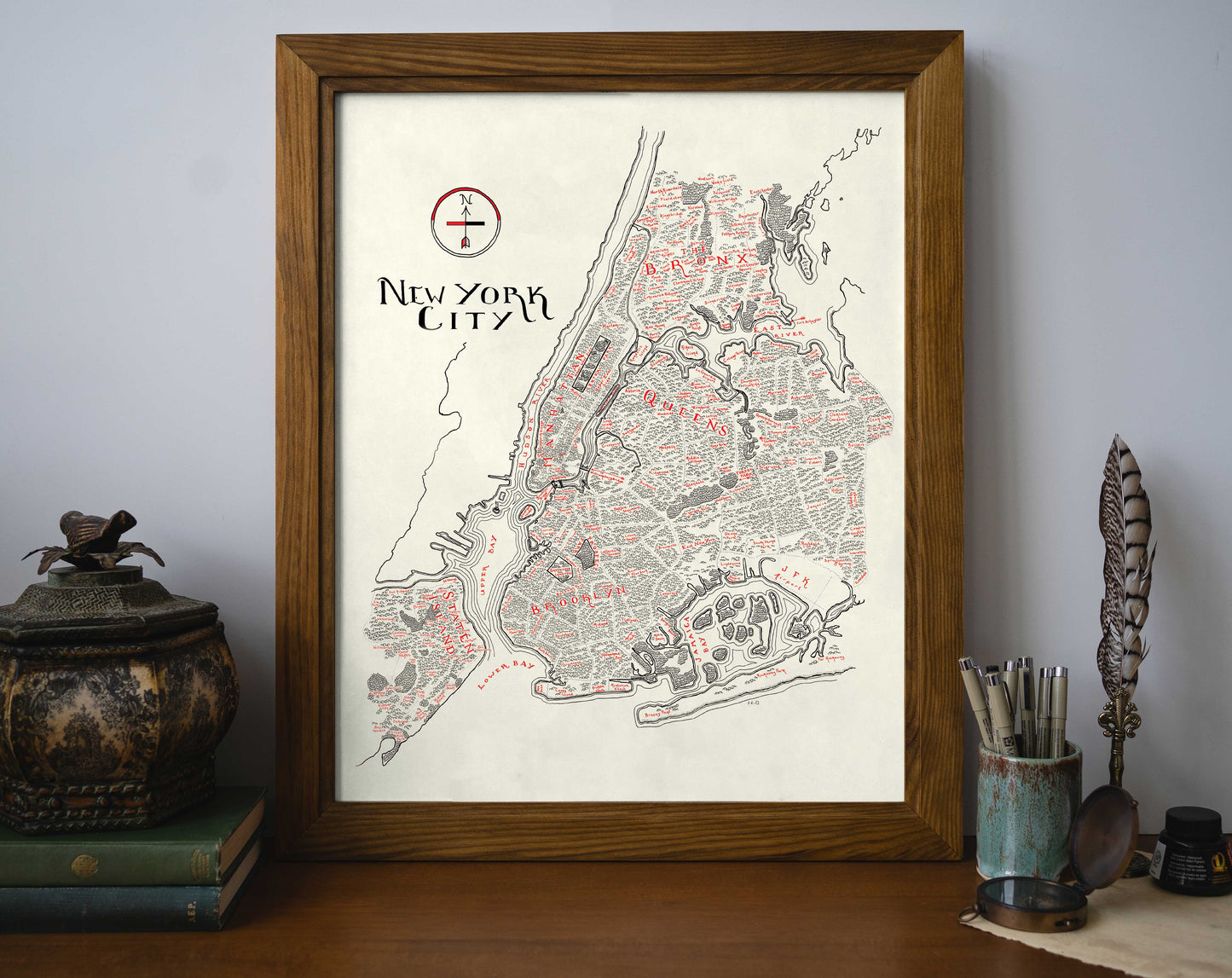 The Lord of Maps New York City Map, featuring bold red and black accents, is framed on a wooden desk beside stacked books, a decorative box, pens in a cup, and a quill holder.
