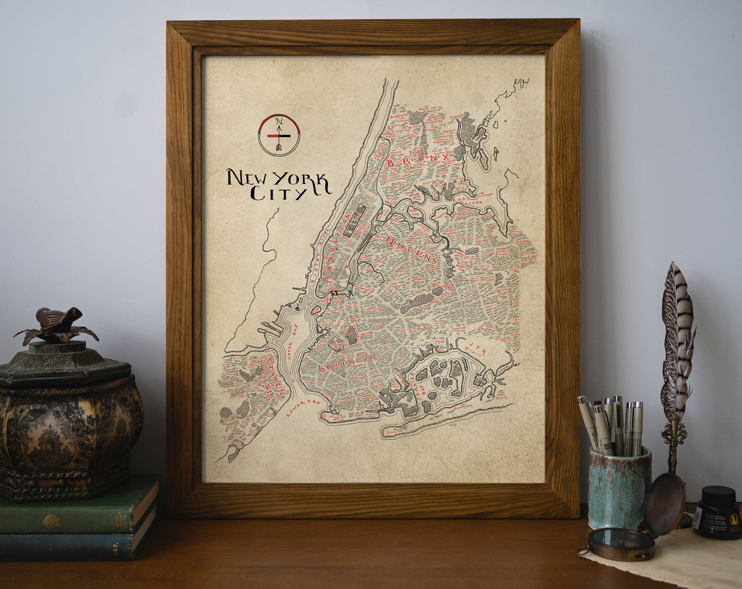 The Lord of Maps New York City Map features black and red detailing on a beige background. Displayed framed on a desk, it sits beside books, a decorative container, a quill, and cups filled with pens and pencils.