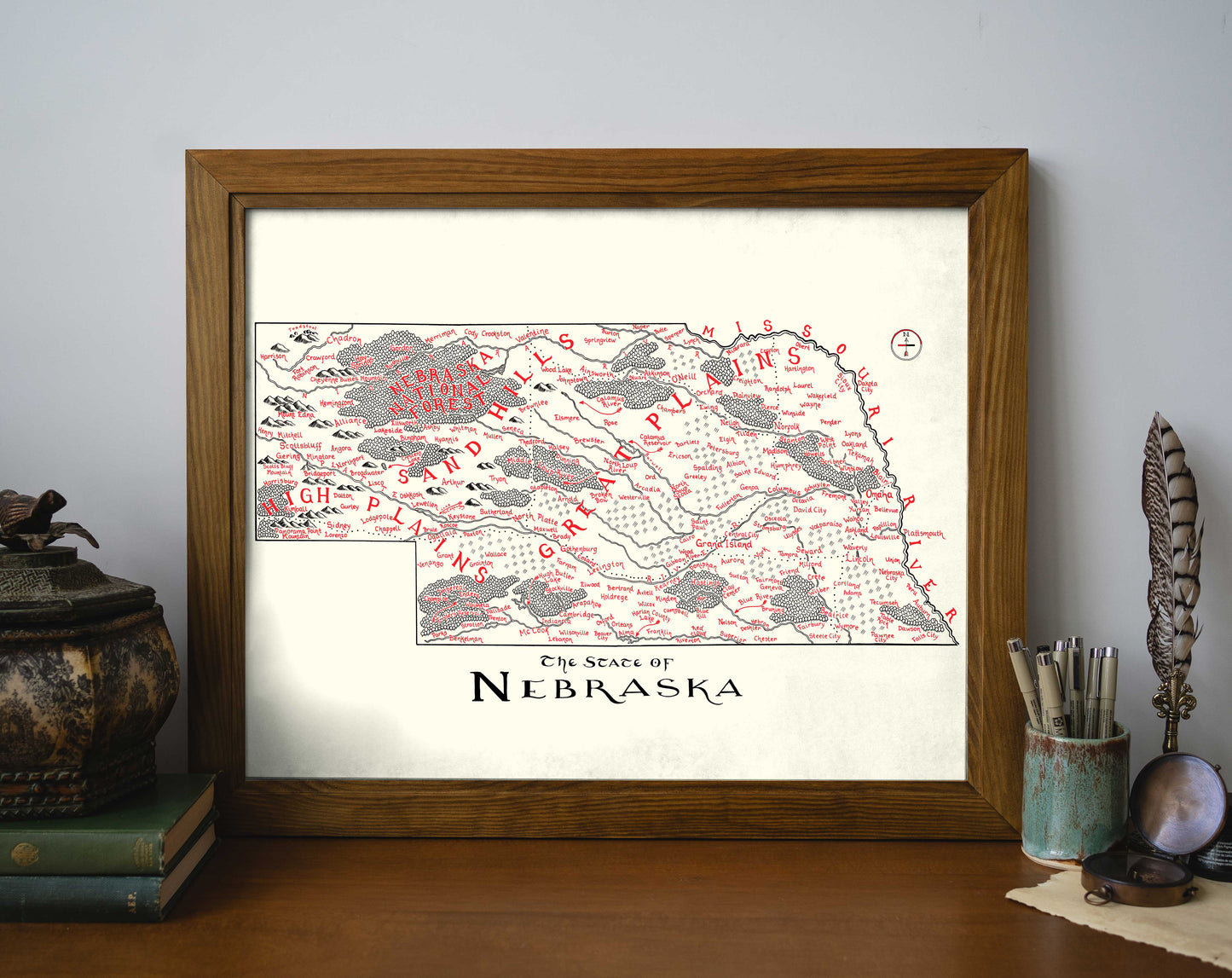 The Lord of Maps Nebraska Map is a framed, hand-drawn wall art print with red and gray markings, labeled "The State of Nebraska," shown on a desk beside books, a plant, and decor items.