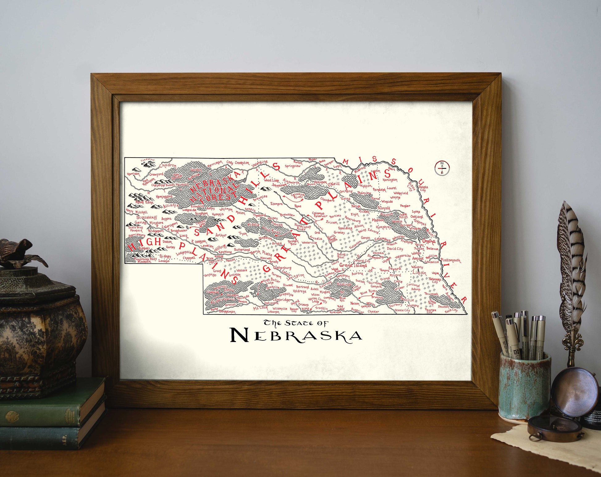 The Lord of Maps Nebraska Map is a framed, hand-drawn wall art print with red and gray markings, labeled "The State of Nebraska," shown on a desk beside books, a plant, and decor items.