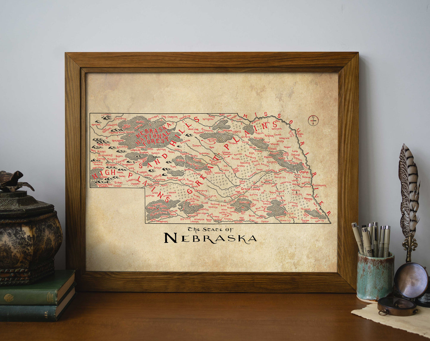 The Nebraska Map by Lord of Maps is a framed, vintage-style print with decorative red markings and labels, shown on a wooden surface among books, a potted plant, and a pen holder.