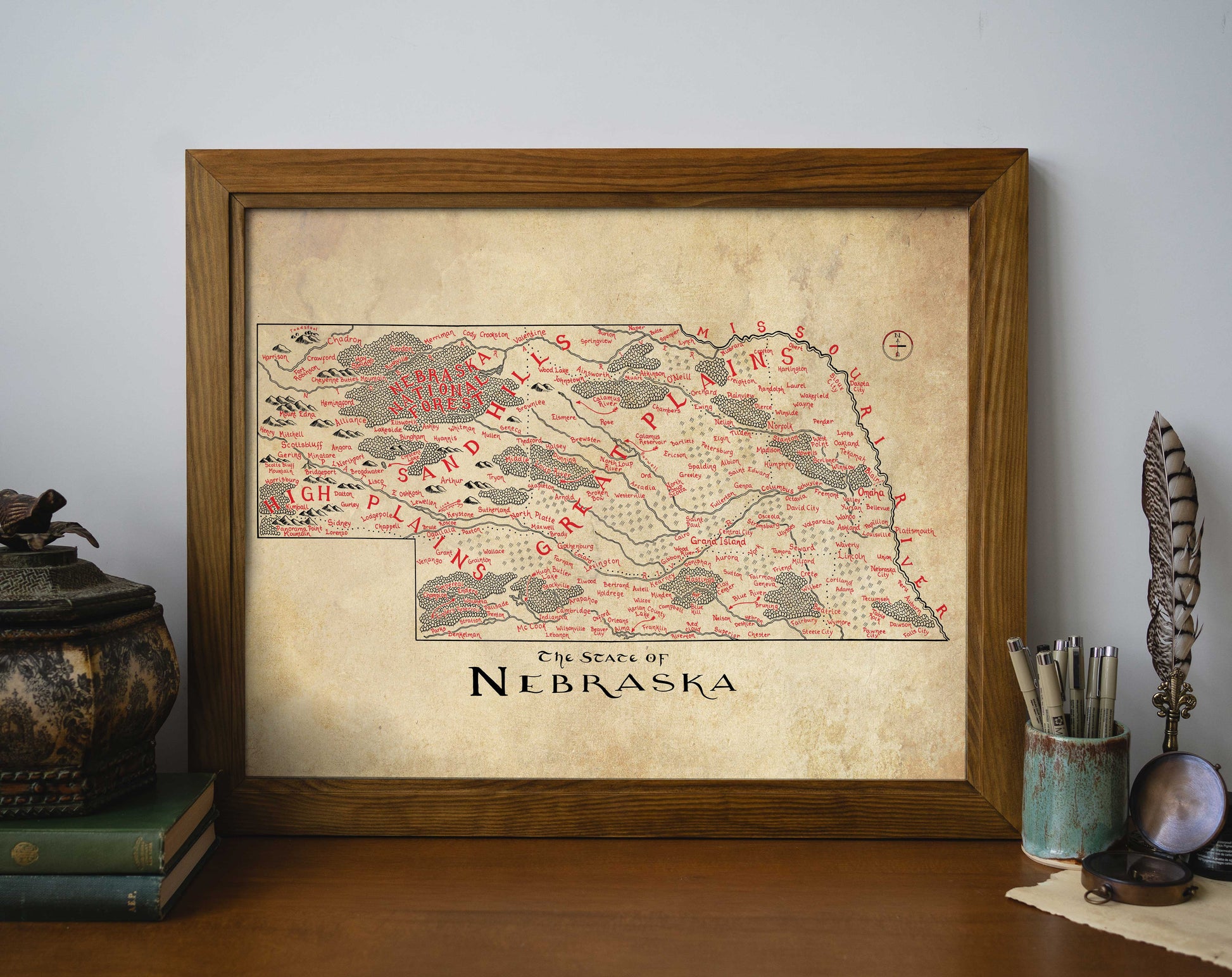 The Nebraska Map by Lord of Maps is a framed, vintage-style print with decorative red markings and labels, shown on a wooden surface among books, a potted plant, and a pen holder.