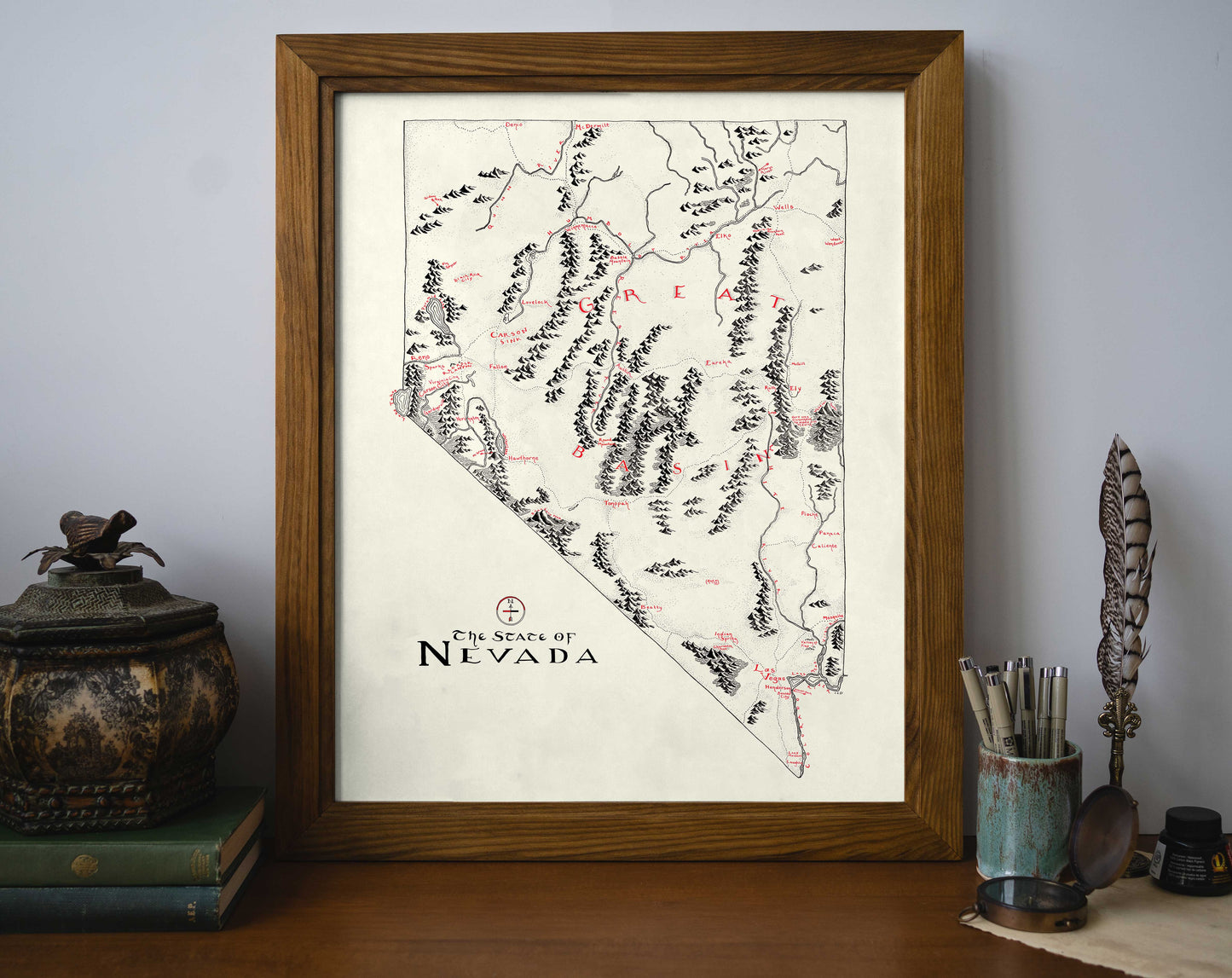 The Nevada Map by Lord of Maps, a framed black-and-white hand-drawn map with red accents, is displayed on a desk among books, a feather quill, a decorative jar, and a container of writing utensils.