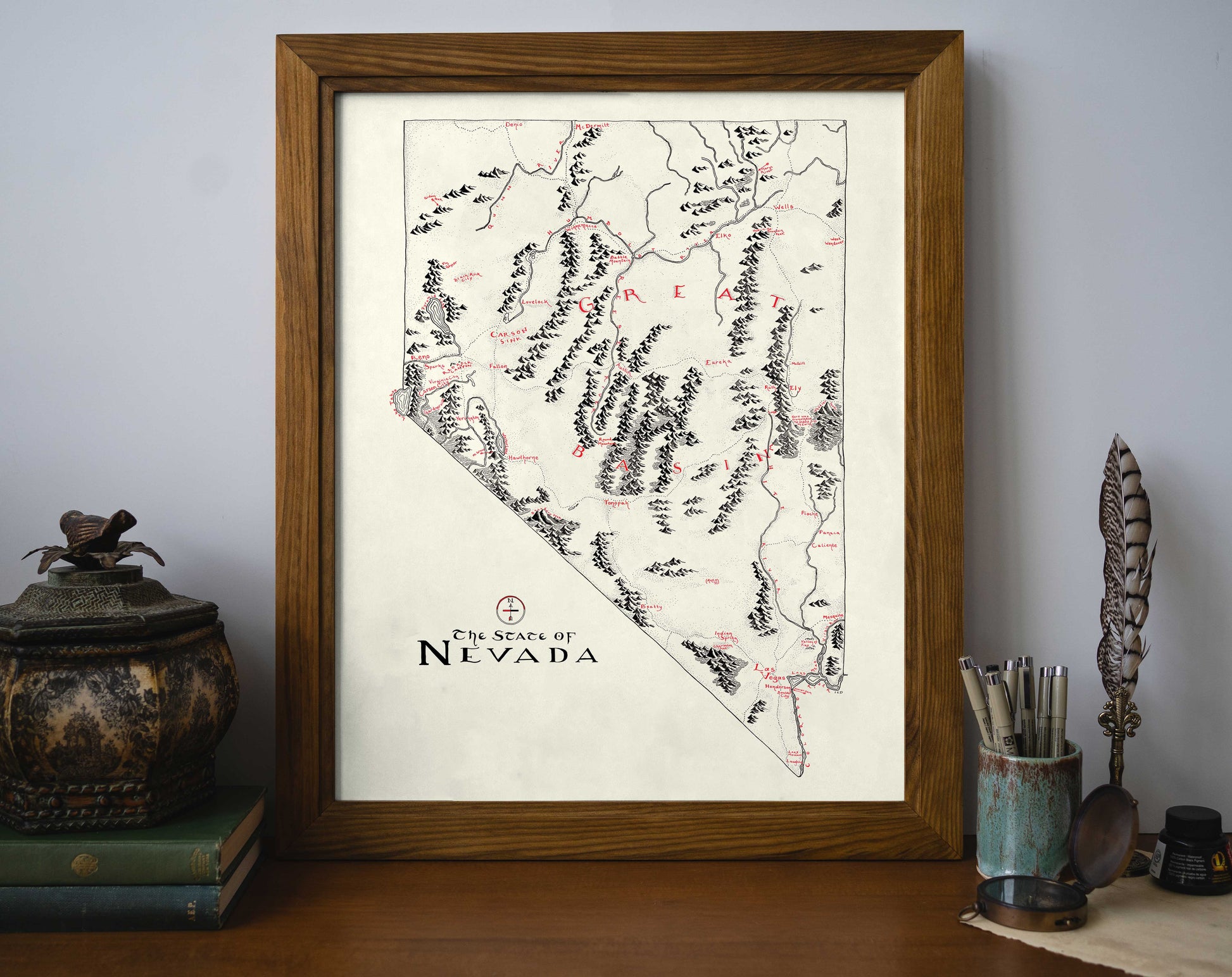 The Nevada Map by Lord of Maps, a framed black-and-white hand-drawn map with red accents, is displayed on a desk among books, a feather quill, a decorative jar, and a container of writing utensils.