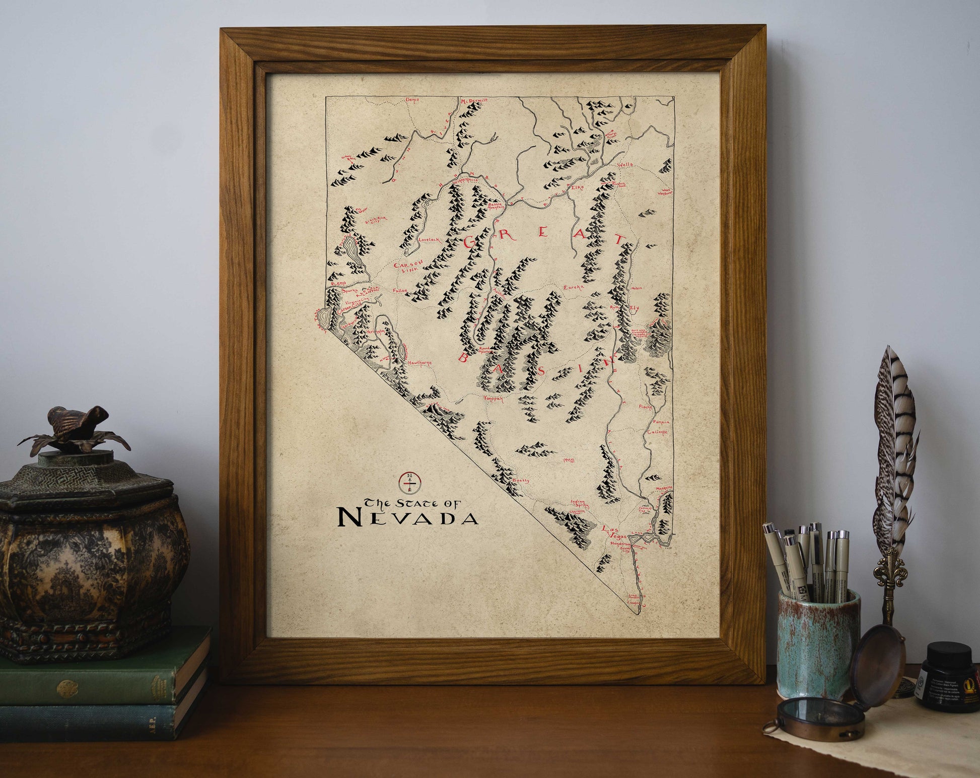 The Nevada Map by Lord of Maps, a vintage-style hand-drawn artwork, is framed and displayed on a desk amid books, decor, and writing tools, creating a cozy and organized workspace against a plain wall.