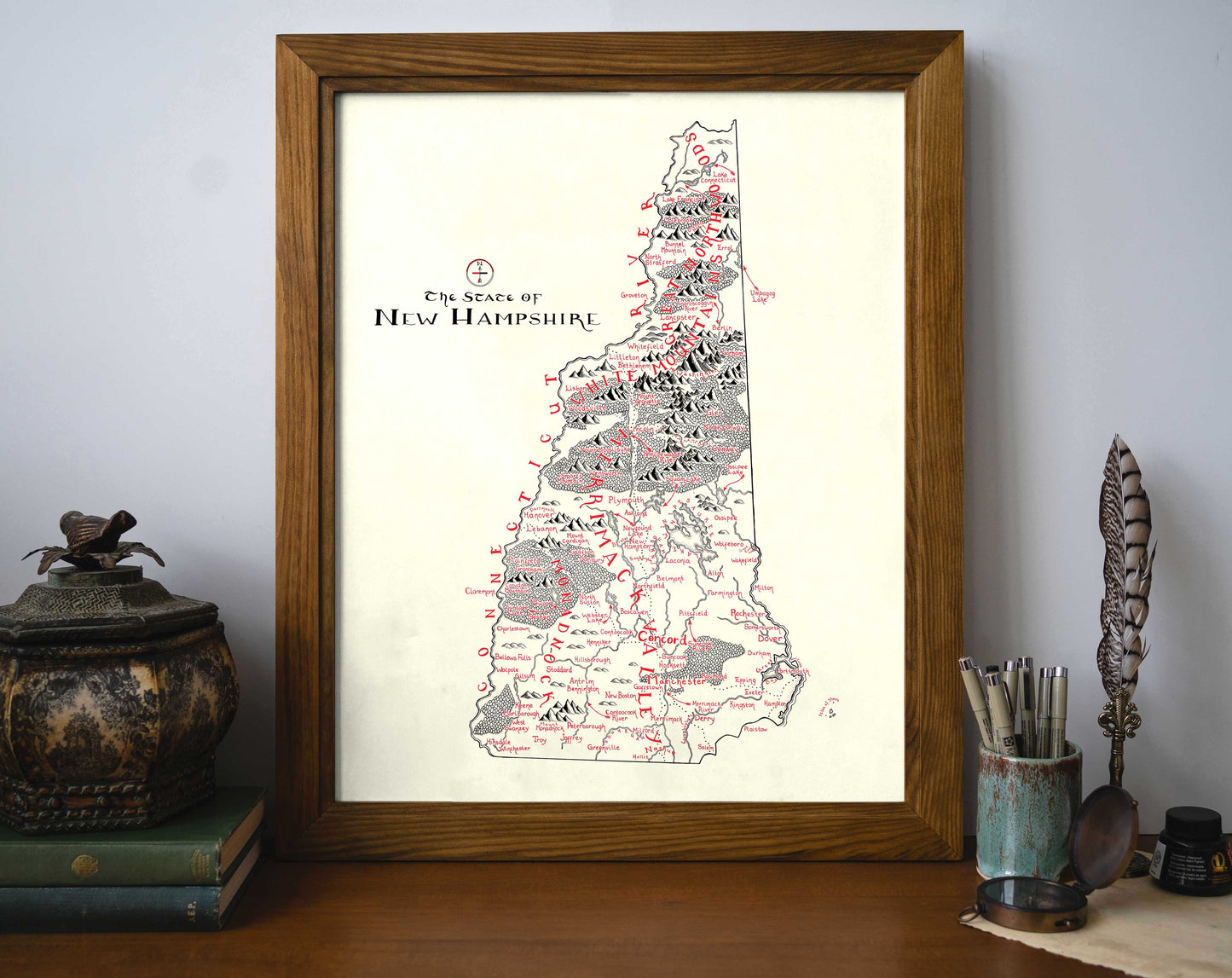 The Lord of Maps New Hampshire Map framed art print sits on a desk against a wall, surrounded by books, an ornate container, and writing tools. This hand-drawn map features handwritten labels and detailed mountain illustrations.