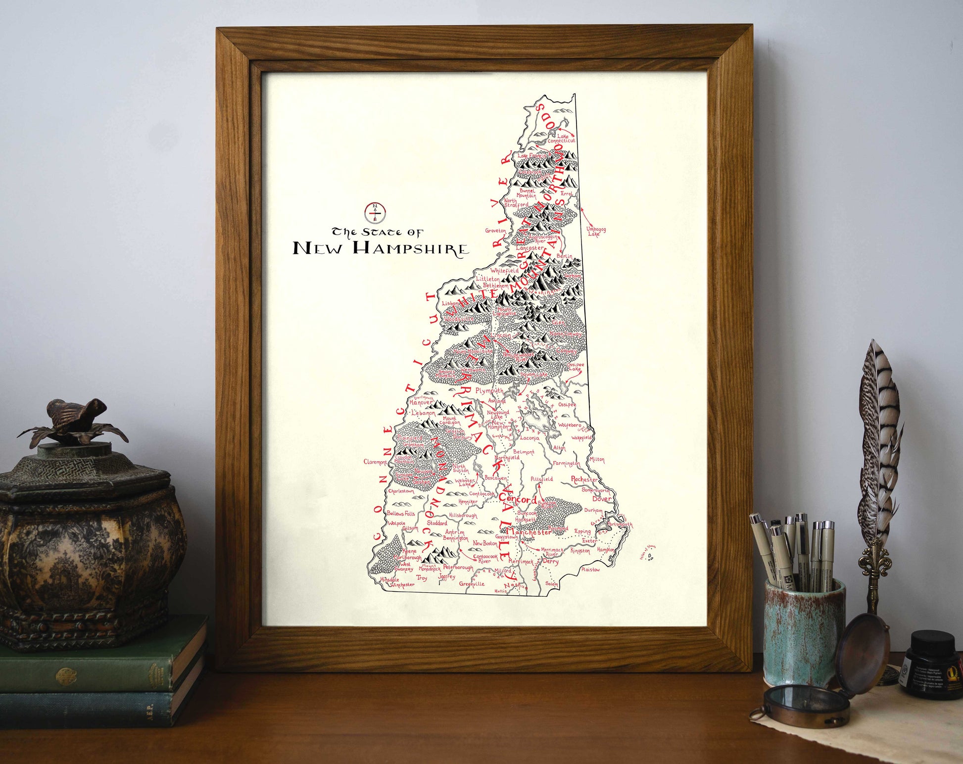 The Lord of Maps New Hampshire Map framed art print sits on a desk against a wall, surrounded by books, an ornate container, and writing tools. This hand-drawn map features handwritten labels and detailed mountain illustrations.