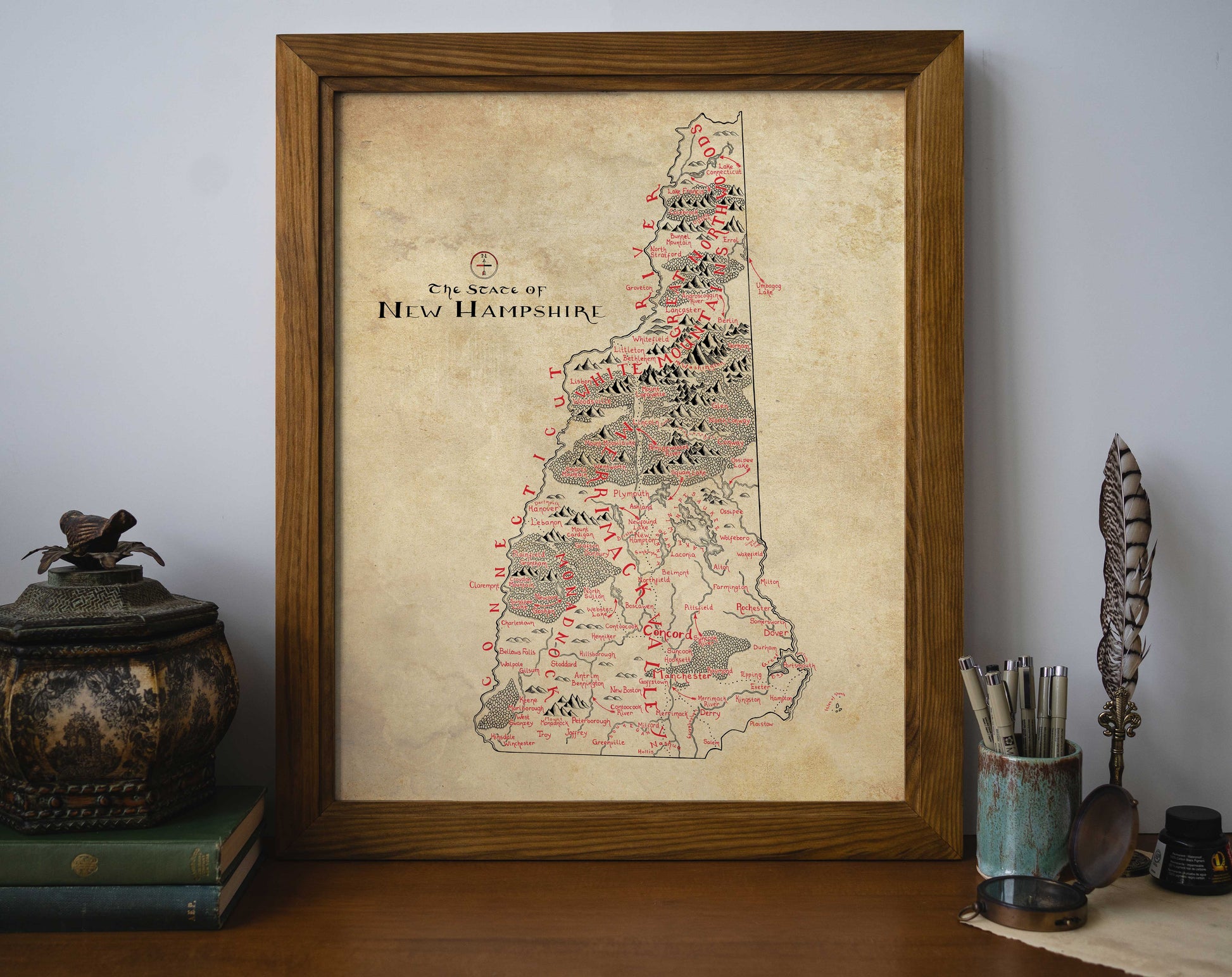 The Lord of Maps New Hampshire Map, a vintage-style hand-drawn print in red and black showing towns and mountains, sits framed on a desk beside books, an ornate jar with a bird figurine, a pen cup with feather quill, and an ink bottle.