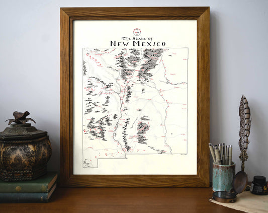 The Lord of Maps New Mexico Map, a vintage-style framed wall art, rests on a desk with a decorative container, stacked books, quill holder, and pens against a plain light wall.