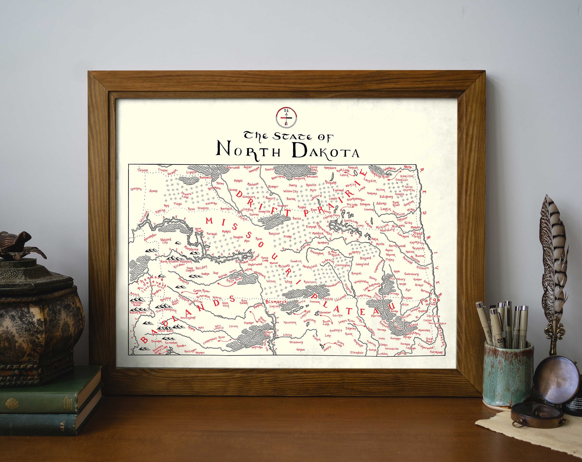 The North Dakota Map by Lord of Maps, featuring hand-drawn red-marked locations, is displayed on a wooden table alongside decorative items, books, and a pen and pencil holder.