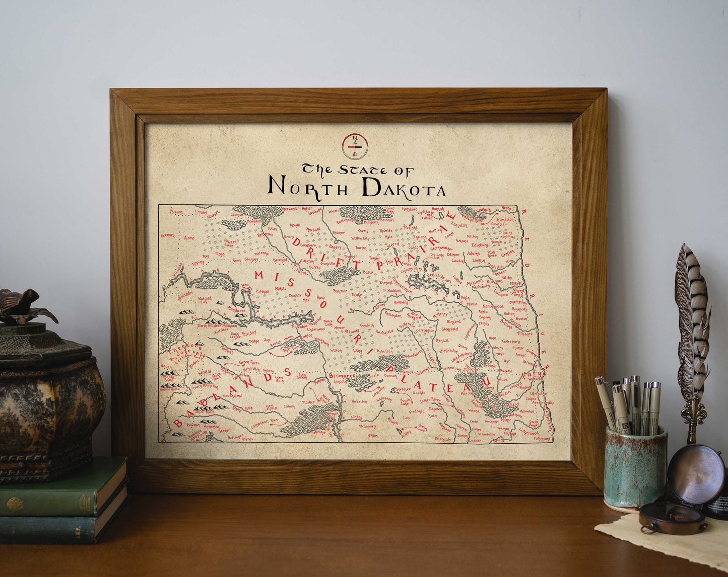 A Lord of Maps North Dakota Map print in a frame sits on a wooden desk, surrounded by books, a pen and pencil holder, and a decorative object.