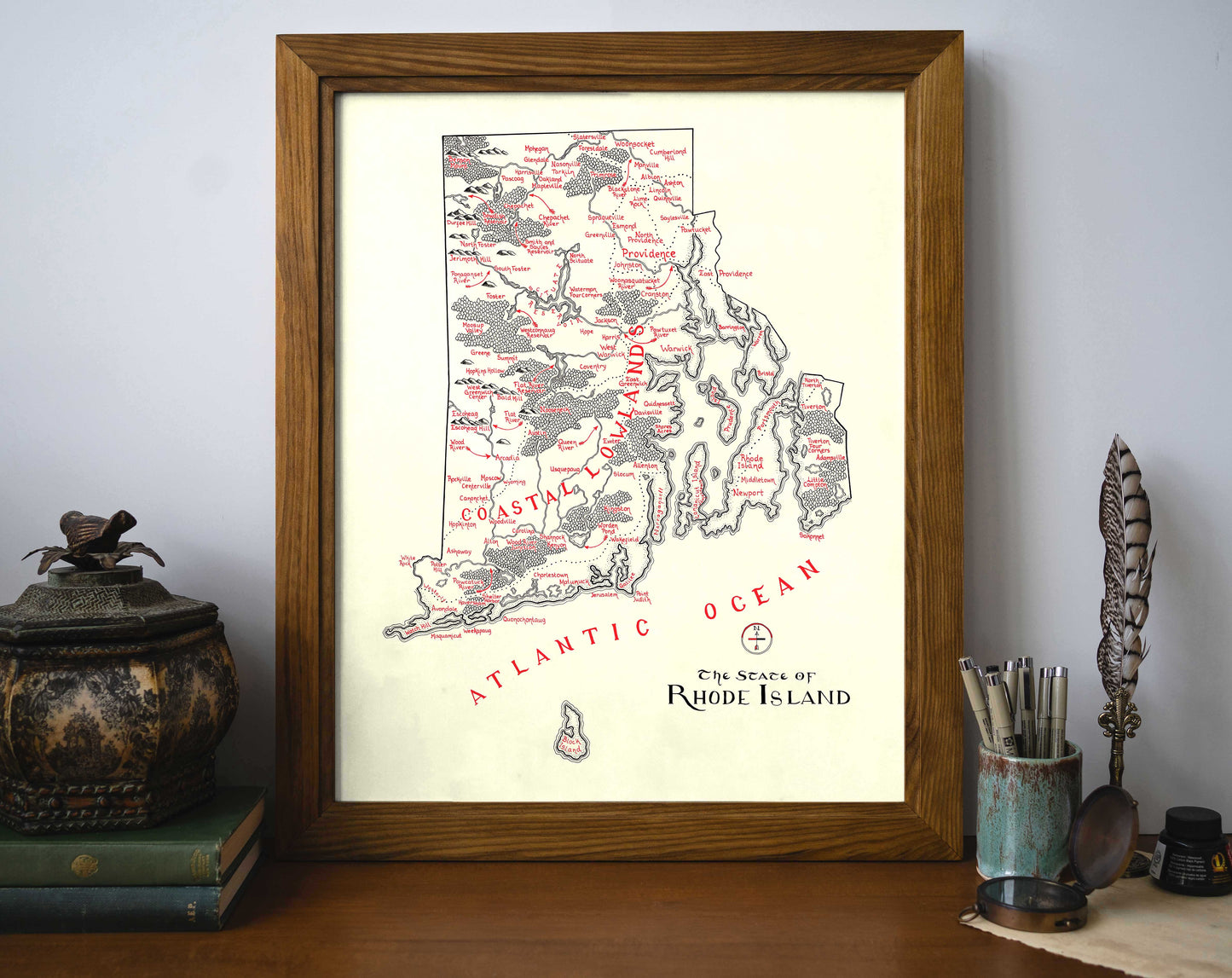 The Rhode Island Map by Lord of Maps, a vintage-style framed wall art labeled with regions, sits on a desk with books, art supplies, and decor. “Atlantic Ocean” and “The State of Rhode Island” appear on the map.