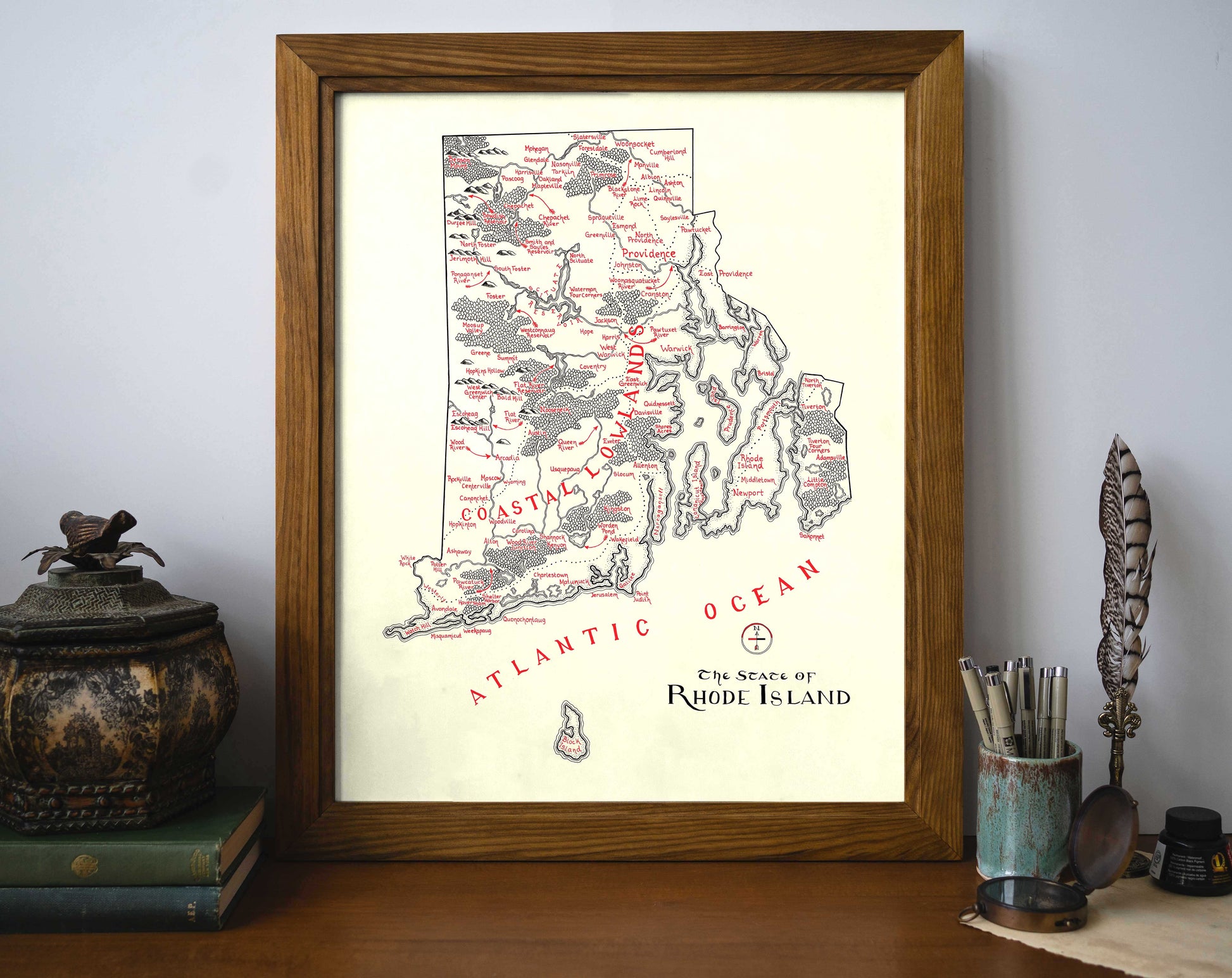 The Rhode Island Map by Lord of Maps, a vintage-style framed wall art labeled with regions, sits on a desk with books, art supplies, and decor. “Atlantic Ocean” and “The State of Rhode Island” appear on the map.