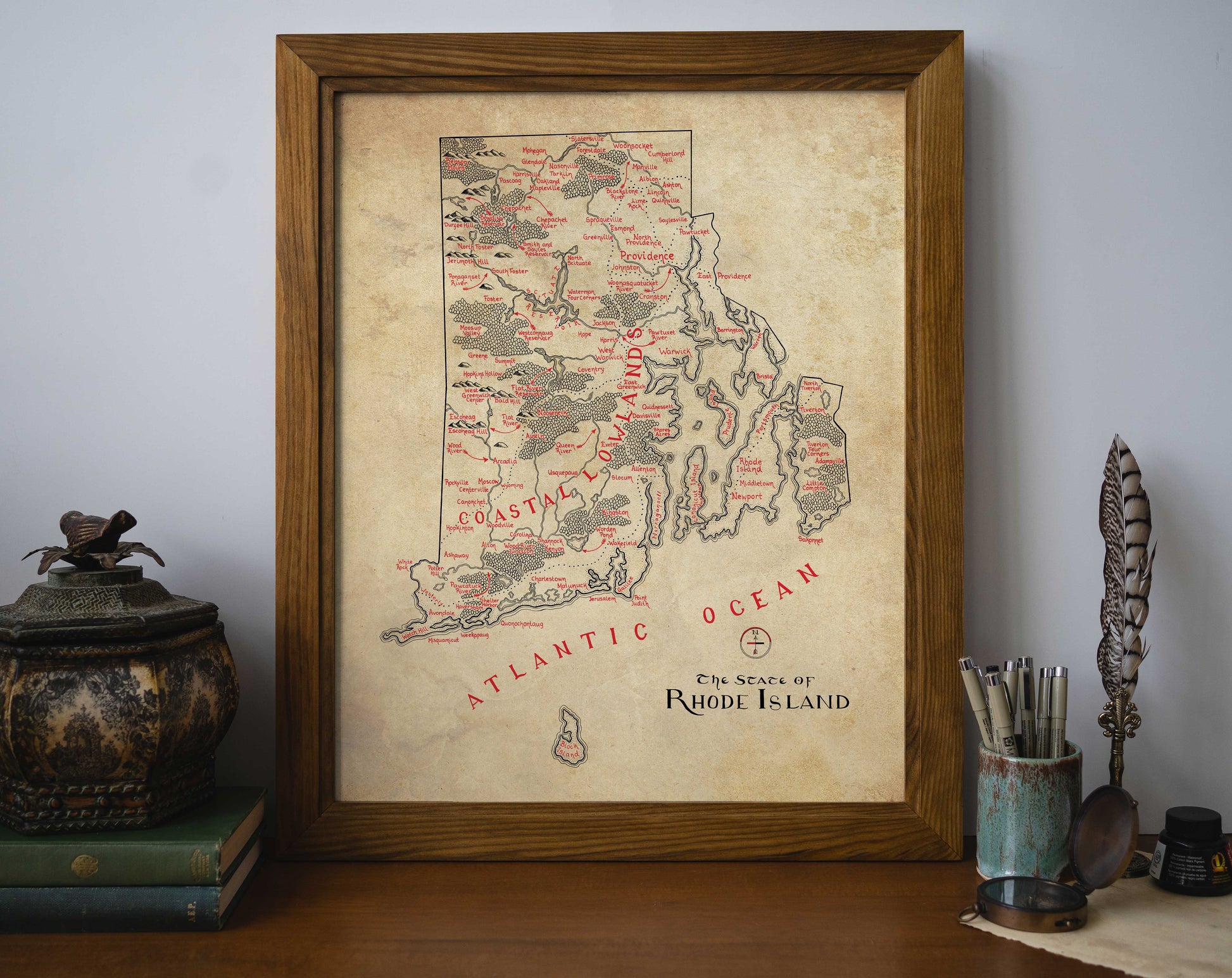 The Lord of Maps Rhode Island Map, a framed vintage-style wall art print, features hand-drawn towns, rivers, and the Atlantic Ocean labeled in red text—perfect for display on your desk alongside books and decor.