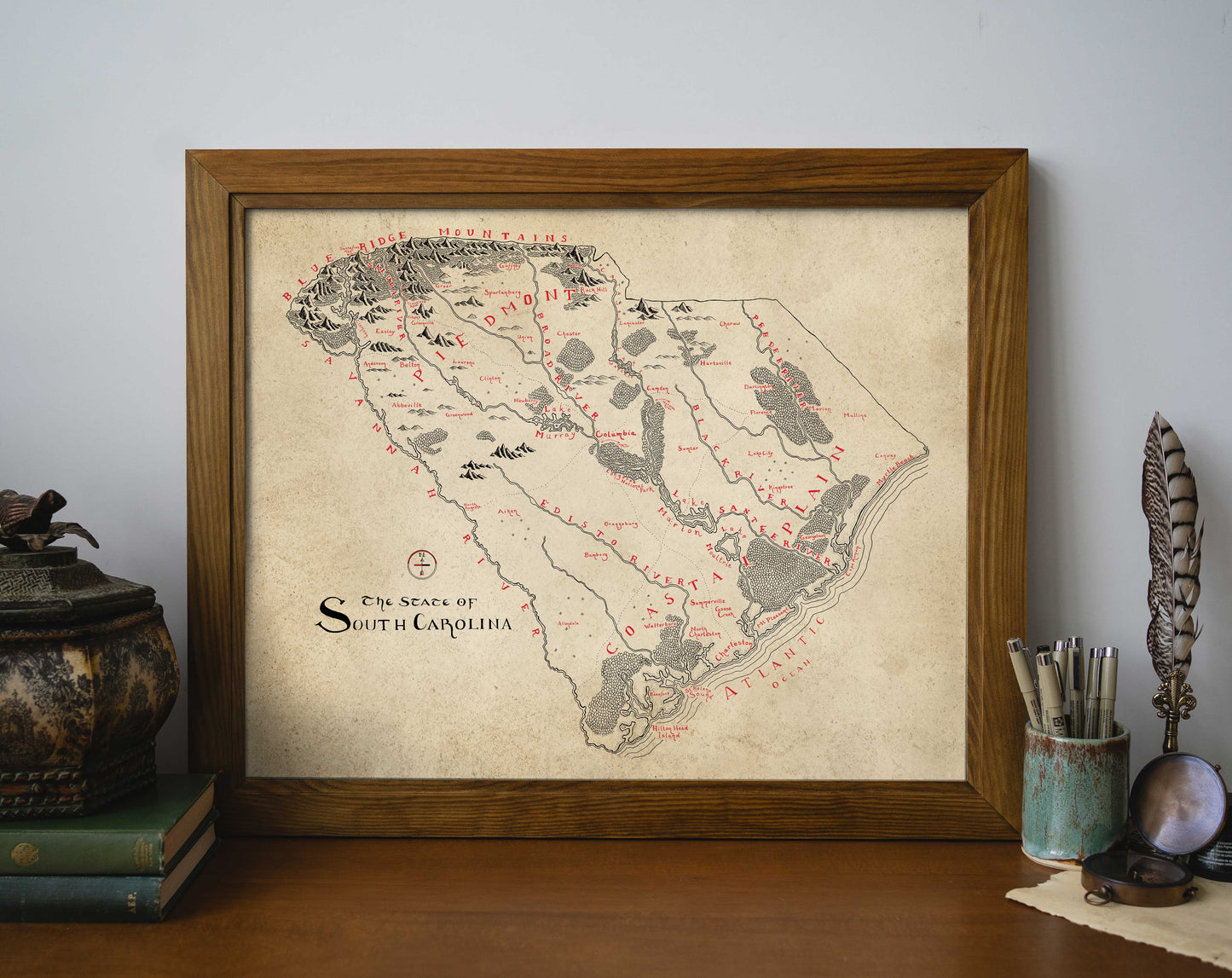 The South Carolina Map by Lord of Maps, a framed vintage-style print, rests on a wooden surface beside stacked books, a decorative container, and a cup with writing utensils and a feather quill.