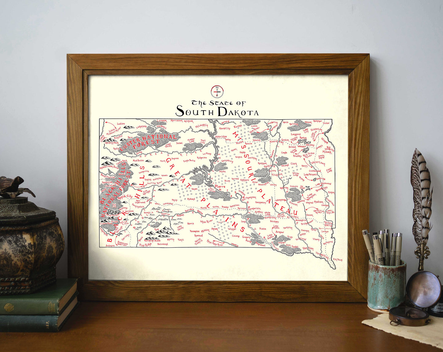 The South Dakota Map by Lord of Maps is a framed hand-drawn print in red and black ink, displayed on wood with books and decor—ideal wall art for any room.
