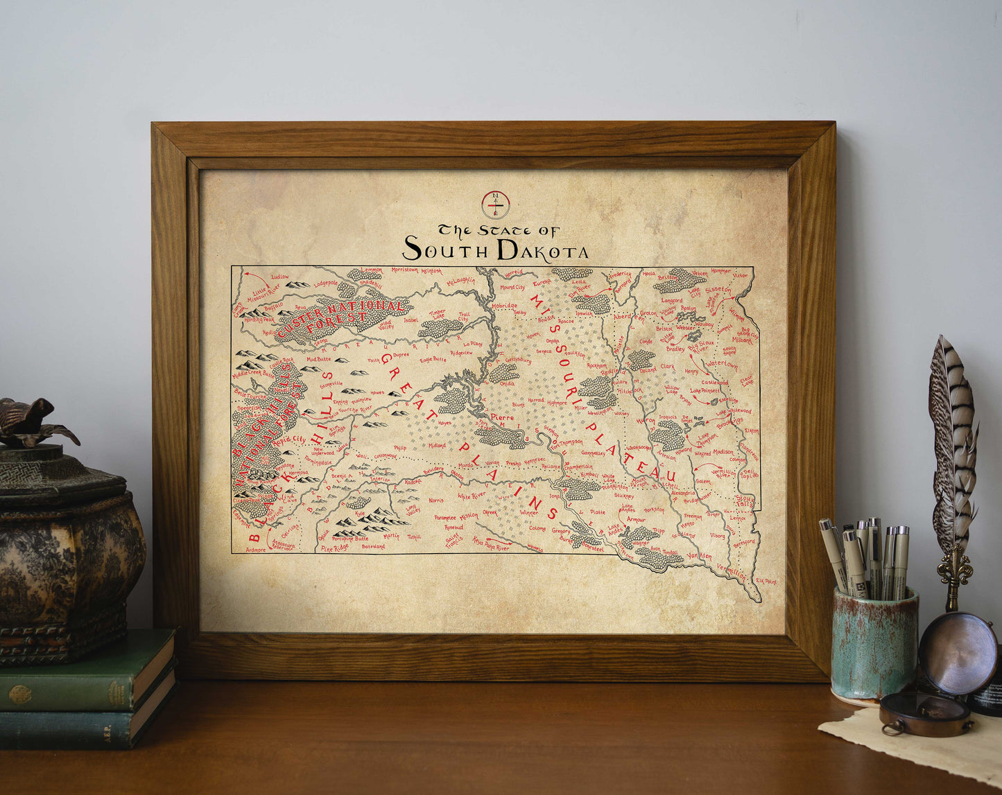 The Lord of Maps South Dakota Map, a framed hand-drawn print with red and black place names and landmarks, sits on a wooden surface surrounded by books, decor, and a feather.