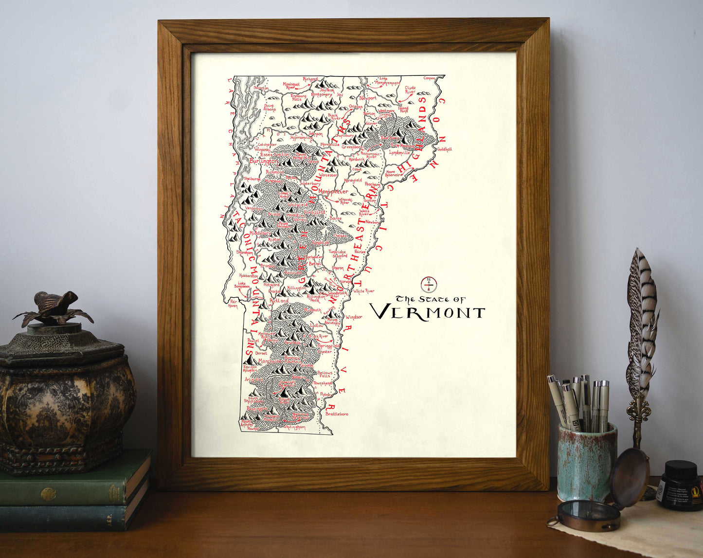 The Lord of Maps Vermont Map is a framed black and red mountain range print, displayed on a shelf beside books, a bird figurine, and a cup of pens and brushes.