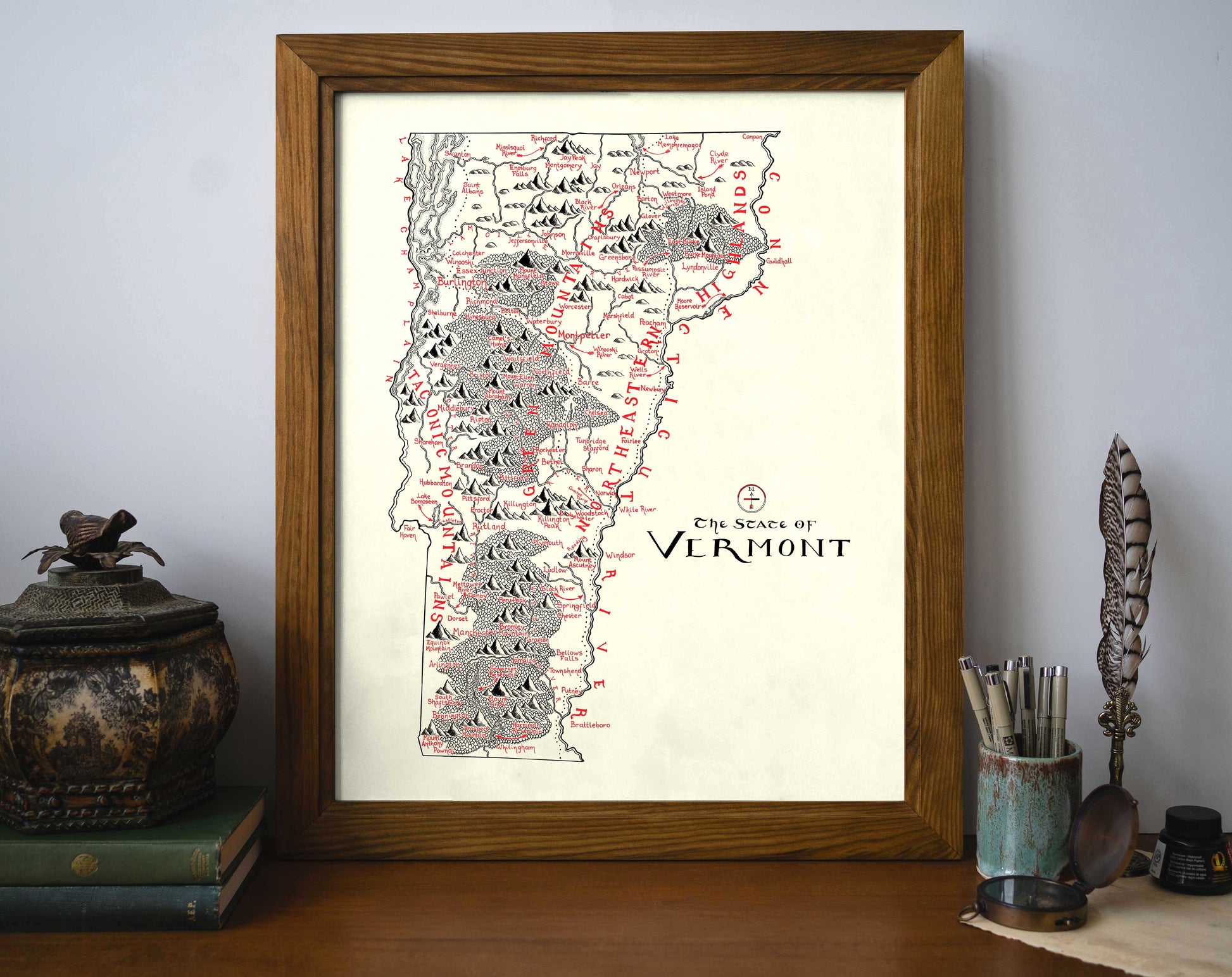 The Lord of Maps Vermont Map is a framed black and red mountain range print, displayed on a shelf beside books, a bird figurine, and a cup of pens and brushes.