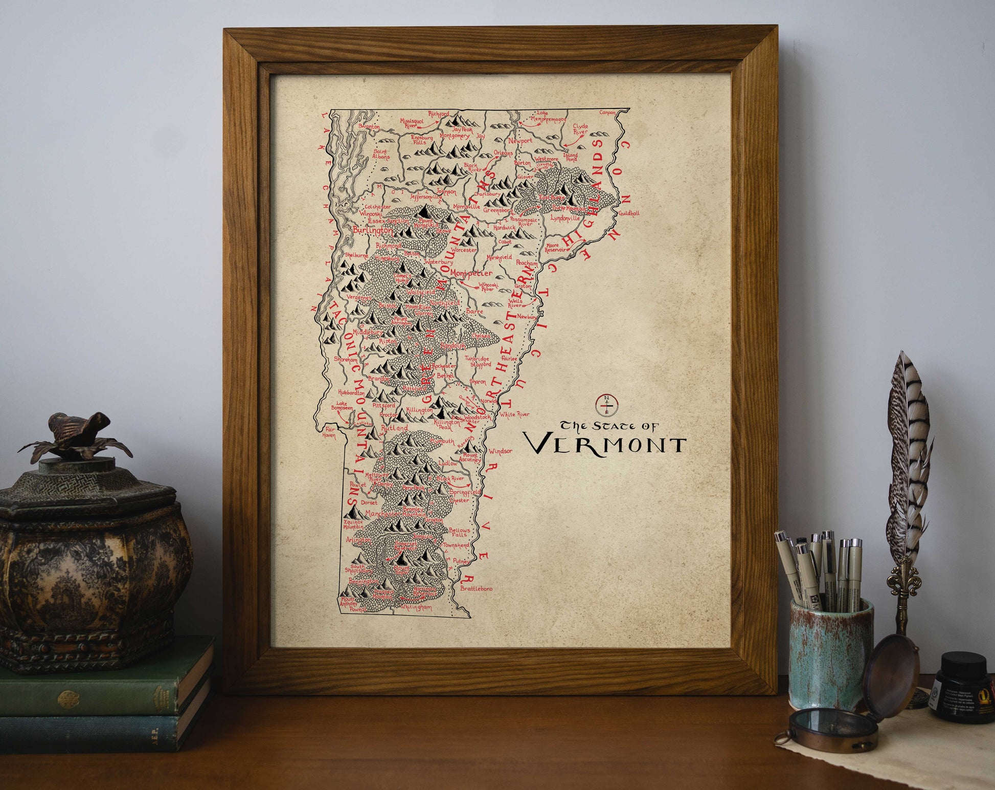 The Vermont Map by Lord of Maps features a hand-drawn, framed print that highlights mountains and towns in black and red ink on a beige background—perfect for display on your desk alongside books and decor.