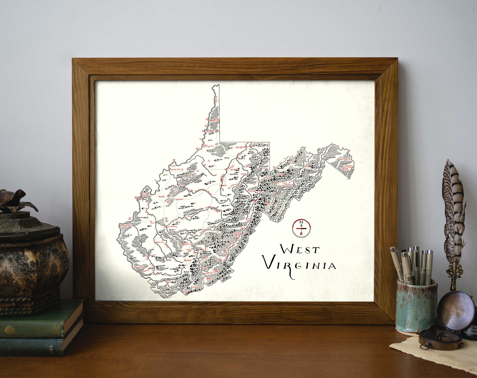 The Lord of Maps West Virginia Map, a framed hand-drawn print with detailed black and red markings and handwritten-style text, is displayed on a desk beside antique books, a decorative container, a quill, and a candlestick.