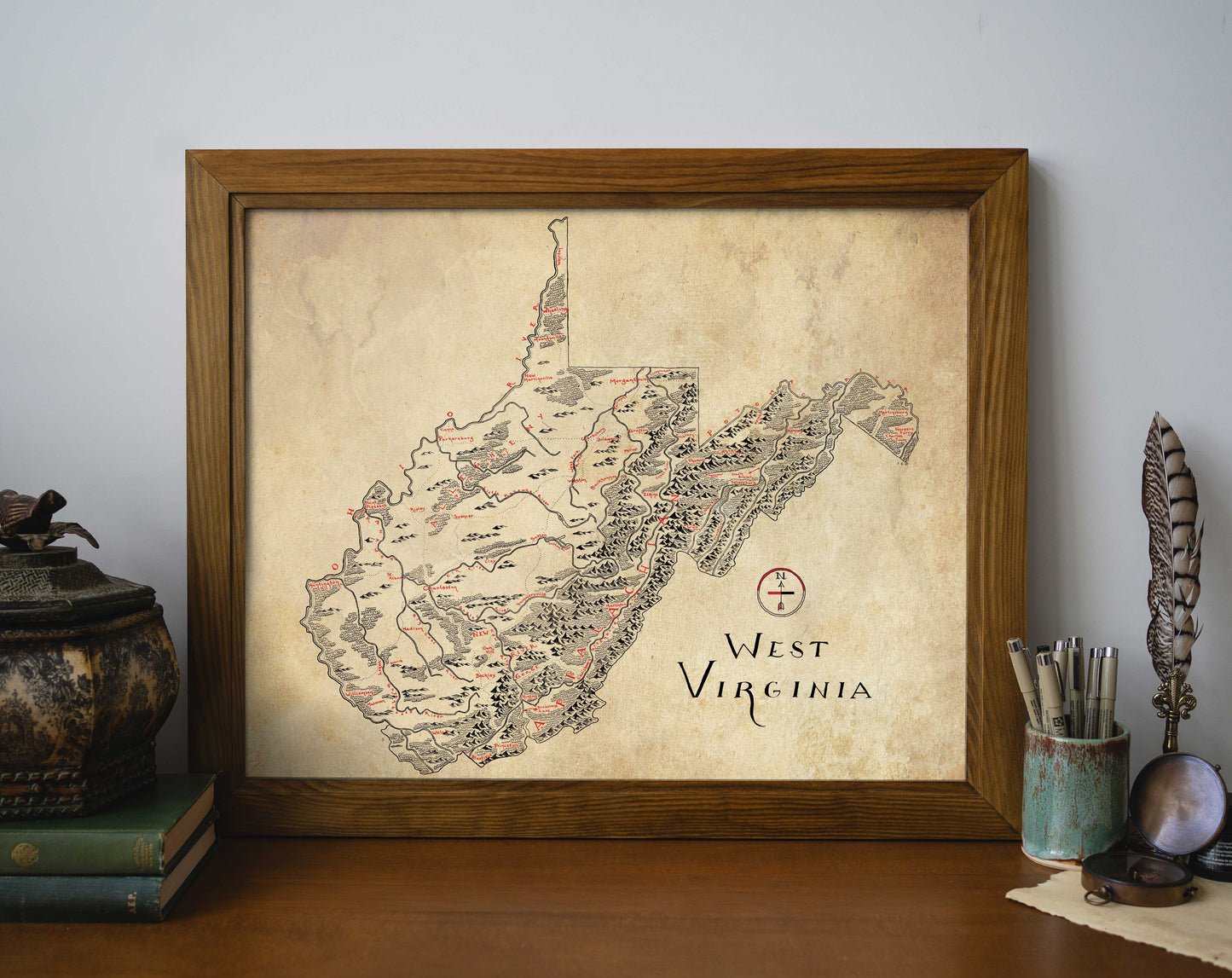 The Lord of Maps West Virginia Map, a framed hand-drawn print with vintage style, features towns and rivers in black and red text. Displayed on a wooden desk with books, a decorative container, feather quill, and jar of pens.
