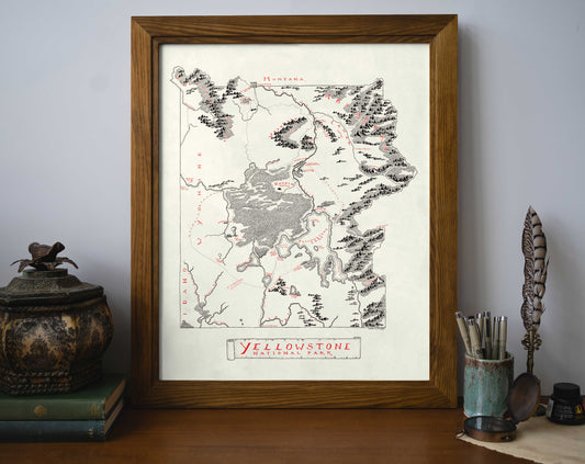 The Lord of Maps Yellowstone National Park Map, a framed vintage-style art print, sits on a desk with books and writing tools. Its hand-drawn design highlights park details and state boundaries in black and red.