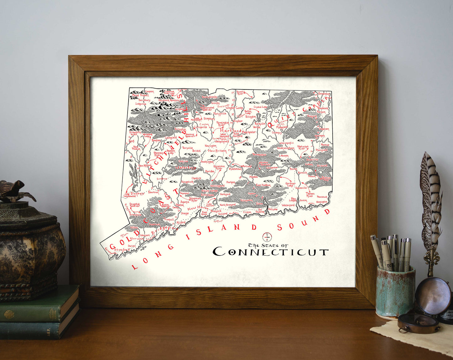 The Connecticut Map by Lord of Maps is a hand-drawn, high-quality print featuring towns, landmarks, and Long Island Sound labeled in red. Shown framed on wood beside books, a feather, and decor.