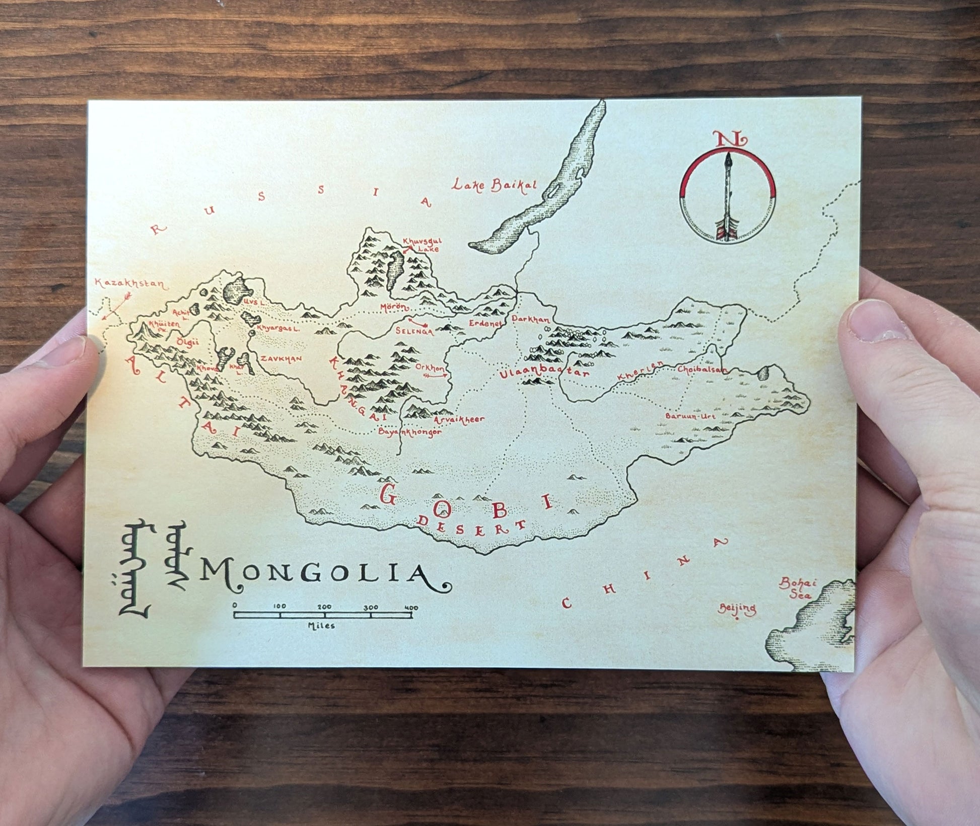A person holds a hand-drawn, vintage-style Lord of Maps Monthly Map Club Mongolia map featuring major cities, rivers, mountains, the Gobi Desert, handwritten labels, and a decorative compass rose—ideal for map enthusiasts.