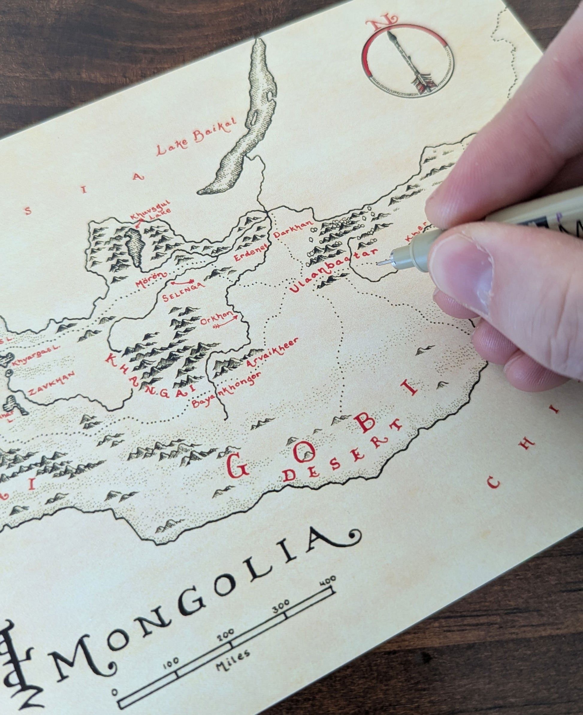 A hand adds details to a vintage-style, hand-drawn map of Mongolia from Lord of Maps’ Monthly Map Club—featuring rivers, mountains, and the Gobi Desert—for fans or anyone wanting a unique map subscription.